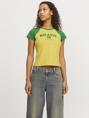 Femme aux cheveux bouclés portant un t-shirt jaune et vert du Brésil et un jean bleu ample, debout devant un fond uni.