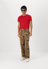 Rode ronde hals t-shirt, olijfgroene camouflage cargo broek met zakken, en witte sneakers, gedragen met zwarte zonnebril tegen een effen achtergrond.