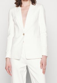 Biała dopasowana marynarka z szpiczastymi klapami, jednolitym złotym zapięciem na jeden guzik oraz bocznymi kieszeniami. Gładka tekstura materiału w połączeniu z dopasowanymi spodniami.