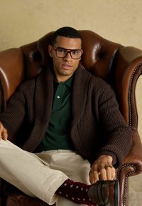 Pruun soonikkardigan roheline polo, beežid püksid ja maroonide mustritega sokid. Istub nahksofas, kandes ruudukujulisi prille.