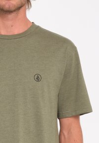 Camiseta de manga corta verde oliva en tejido suave, con un pequeño logo negro en la parte izquierda del pecho, cuello redondo y corte estándar.