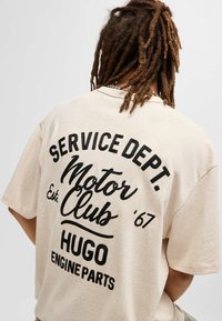 Beige katoenen T-shirt met een relaxed fit, voorzien van een zwarte opdruk: "SERVICE DEPT. Motor Club '67 HUGO ENGINE PARTS" op de achterkant.