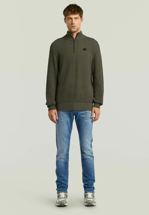 Man staat en draagt een groene gestructureerde trui met een kwart rits, vervaagde blauwe spijkerbroek en grijze en witte sneakers op een effen lichte achtergrond.
