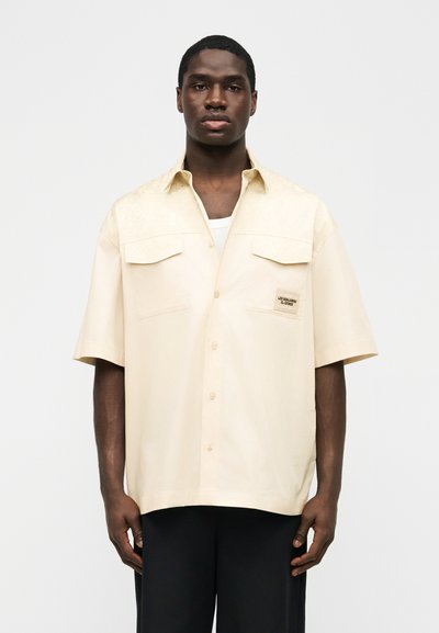 Homme portant une chemise crème à manches courtes avec deux poches poitrine et un pantalon noir, debout devant un fond uni.