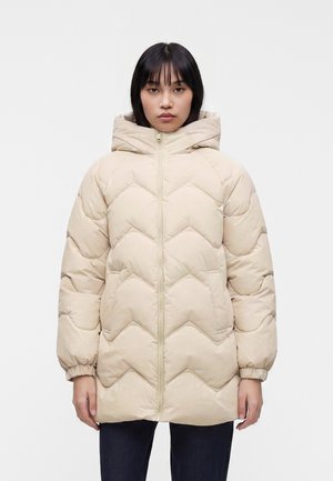 Femme portant une doudoune matelassée beige avec capuche, fermeture éclair à l'avant et poignets élastiques, debout devant un fond blanc uni.
