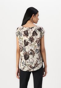 Blouse florale crème avec des motifs de feuilles brunes, manches courtes et détail de fermeture éclair au dos. Portée avec un pantalon noir. Texture de tissu lisse.