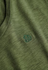 Gros plan sur un tissu vert avec un petit logo vert brodé près de l'encolure sur un vêtement à la texture douce.