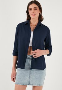 Camicia blu navy con bottoni e maniche lunghe arrotolate fino ai gomiti, realizzata in tessuto leggero, abbinata a una mini gonna in denim chiaro.