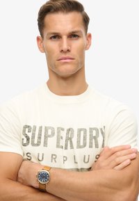Ung mand med lysebrunt hår iført en hvid "Superdry Surplus" t-shirt og et ur med blå urskive og tan læderrem.