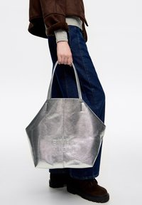 Osoba trzymająca dużą metaliczną torbę w kolorze srebrnym z wytłoczonym napisem "BIMBA Y LOLA", ubrana w niebieskie dżinsy, brązowe buty i brązową kurtkę.