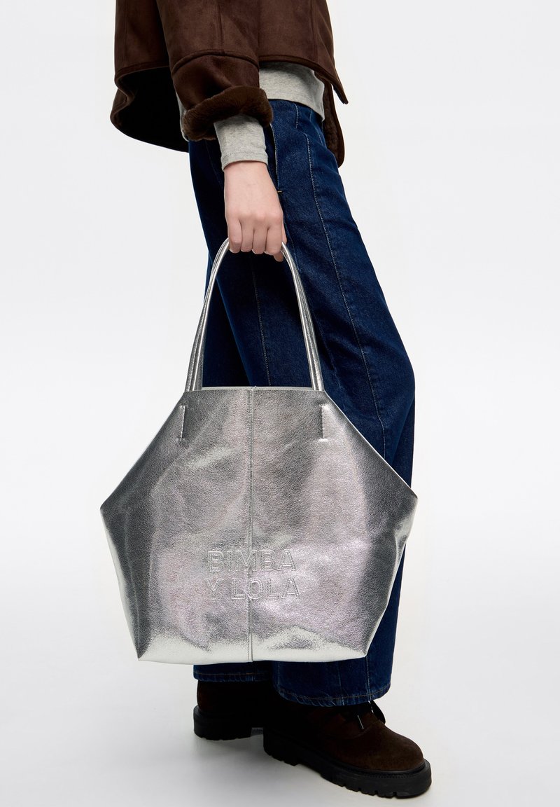 Osoba trzymająca dużą metaliczną torbę w kolorze srebrnym z wytłoczonym napisem "BIMBA Y LOLA", ubrana w niebieskie dżinsy, brązowe buty i brązową kurtkę.