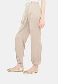 Pantaloni beige, leggeri, con vita elastica, pieghe e orli a risvolto, abbinati a ballerine piatte oro metallico con un accento decorativo.
