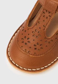 Καφέ δερμάτινο παπούτσι με ανθισμένα σχέδια διάτρησης, μπεζ ραφές κατά μήκος της άκρης και στρογγυλεμένη μύτη σε λευκό φόντο.