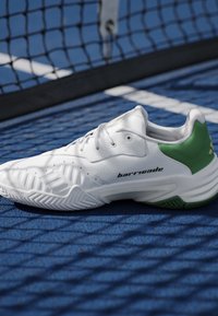 Scarpa da tennis bianca con dettaglio verde sul tallone e testo "barricade", appoggiata su una superficie del campo blu vicino a una rete nera.