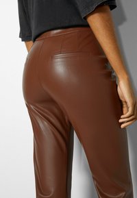 Pantaloni in pelle marrone con una superficie liscia, dotati di tasche posteriori e una vestibilità su misura. L'immagine mostra i dettagli posteriori e laterali.