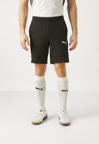 Schwarze Sportshorts mit elastischem Bund und weißem Puma-Logo, kombiniert mit weißen knielangen Socken mit schwarzem Logo und weißen Turnschuhen.
