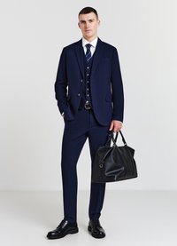 Abito blu navy con un design aderente, caratterizzato da un blazer a tre bottini e pantaloni abbinati. Borsa a tracolla in pelle nera, scarpe nere lucide.