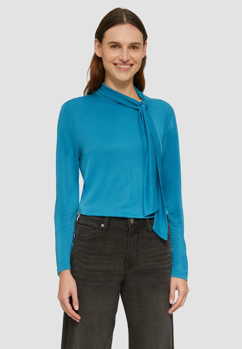 Blauer langärmeliger Oberteil mit geknotetem Halstuchdetail, aus leichtem Stoff hergestellt. Kombiniert mit dunkelgrauen Jeans, minimalistischem Design.