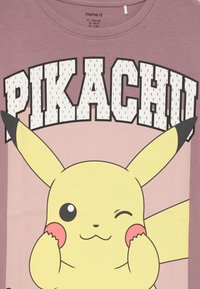 Mirksintis Pikachu su pakeltomis rankomis, liesdamas skruostus po dideliu "PIKACHU" tekstu ant mauvinės spalvos marškinėlių.