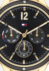 Montre analogique ronde avec un cadran noir, des index et des aiguilles dorés. Comprend trois sous-cadrans et un petit accent en cristal à 12 heures.