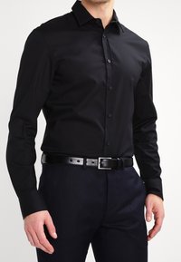 Chemise noire à manches longues avec un col structuré, une patte de boutonnage à l'avant et un tissu lisse. Portée avec une ceinture en cuir noir et un pantalon bleu marine.