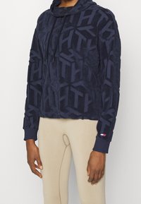 Mörkblå kort hoodie i mjukt, texturerat tyg. Har ett genomgående geometriskt mönster, justerbar dragsko på huvan och ribbade ärmslut.