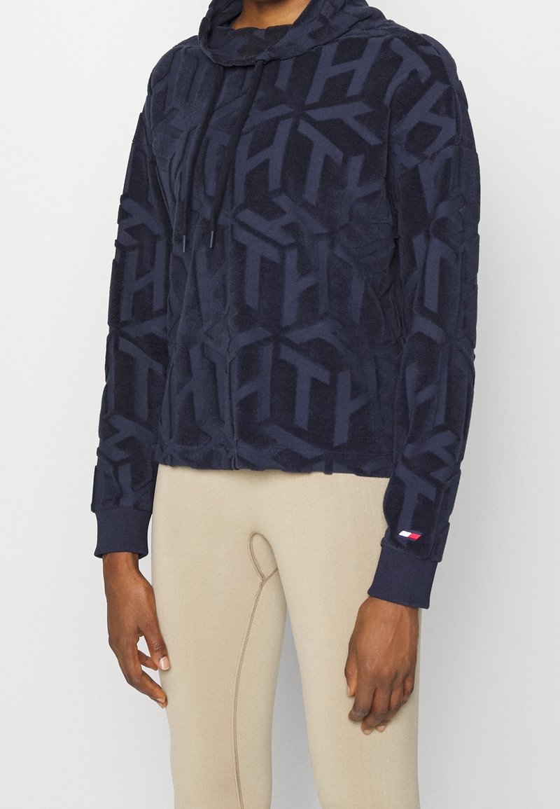 Mörkblå kort hoodie i mjukt, texturerat tyg. Har ett genomgående geometriskt mönster, justerbar dragsko på huvan och ribbade ärmslut.