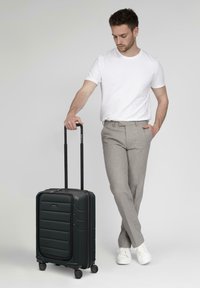 Valigia nera rigida con una parte anteriore texture, maniglia estensibile e quattro ruote, posizionata accanto a una persona in abbigliamento casual.
