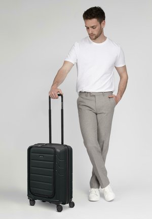 Valigia nera rigida con una parte anteriore texture, maniglia estensibile e quattro ruote, posizionata accanto a una persona in abbigliamento casual.