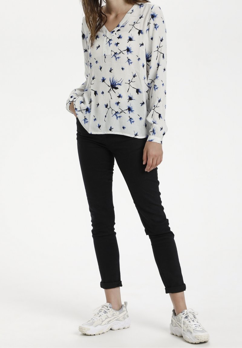 Femme portant un chemisier blanc à manches longues avec un imprimé floral bleu, un pantalon noir moulant retroussé aux chevilles et des baskets blanches, debout avec une main dans la poche.