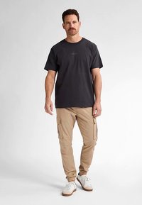 Černé bavlněné tričko s krátkými rukávy, doplněné béžovými kapsáčovými kalhotami se bočními kapsami a bílými teniskami s hnědými podrážkami.