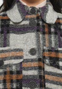 Geruit wollen jas met grijze, oranje en paarse strepen, voorzien van een afgeronde kraag en vier zwarte knopen aan de voorkant.