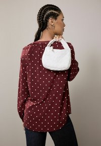 Bolso tejido blanco con forma redondeada y asa anudada, sostenido contra una blusa burdeos de lunares. Textura de tela suave visible.
