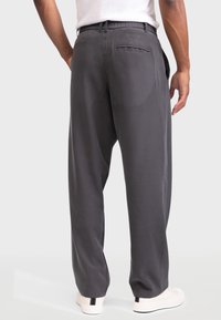 Hombre con pantalones sueltos gris oscuro y zapatillas blancas, de pie con la espalda a la cámara sobre un fondo blanco.