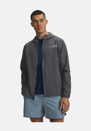 Jonge man met een grijze Under Armour hoodie over een marineblauw shirt en lichtblauwe sportshort, staand met ontspannen handen.