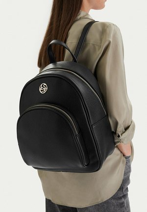 Žena s černým koženým batohem se dvěma kapsami na zip a malým zlatým kruhovým logem na horní přední kapse.
