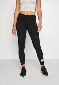 Black leggings made of stretchy material, featuring a wide waistband and a white logo print on the lower leg. Designed for athletic use.