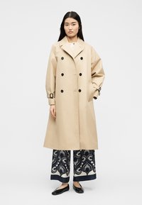 Γυναίκα που φοράει μπεζ διπλόμπουκο μπουφάν με μανσέτες με πόρπες, παντελόνια με φαρδιά πόδια και σχέδια, και μαύρα μπαλαρίνες, στέκεται μπροστά σε λευκό φόντο.