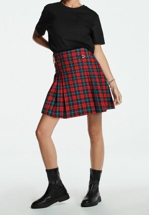 Kobieta ubrana w czarny t-shirt, czerwoną i zieloną plisowaną spódnicę w kratę oraz czarne botki, pozująca z jedną ręką na biodrze.