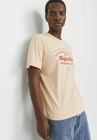 Licht beige t-shirt met korte mouwen en een ronde halslijn, met rode tekstgraphics die "Surfer's Club" op de borst weergeven.