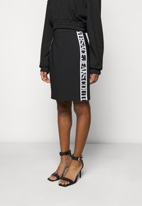 Kvinna i en svart kjol med vit vertikal text "Versace Jeans Couture" på sidan, tillsammans med svarta högklackade sandaler med ankelrem.