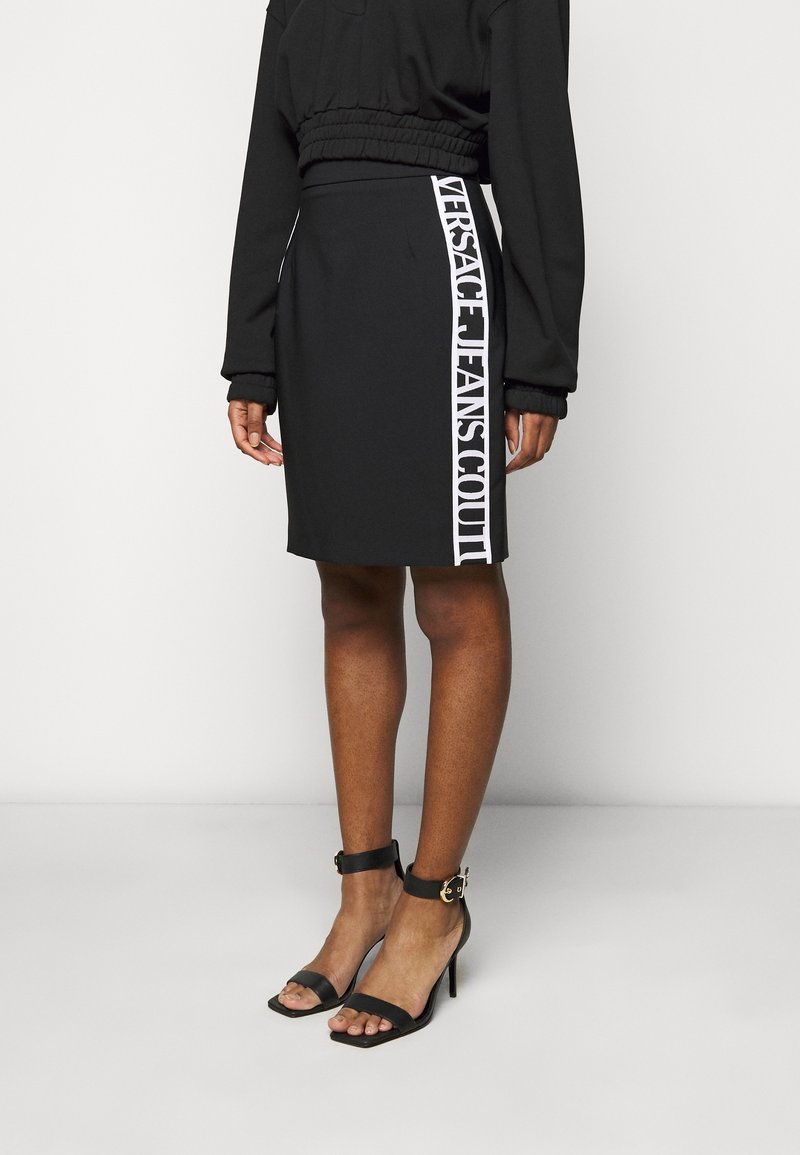 Kvinna i en svart kjol med vit vertikal text "Versace Jeans Couture" på sidan, tillsammans med svarta högklackade sandaler med ankelrem.