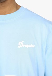 Világoskék pamut pulóver, kerek nyakkivágással és fehér "Dropsize" logóval a mellrészen. Simább textúra és laza fazon jellemzi.