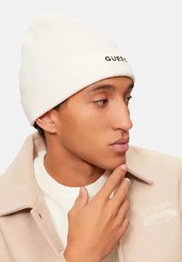 Beanie bianco con logo "GUESS", indossato da un modello in giacca beige su un maglione crema. Tessuto liscio con una vestibilità aderente.