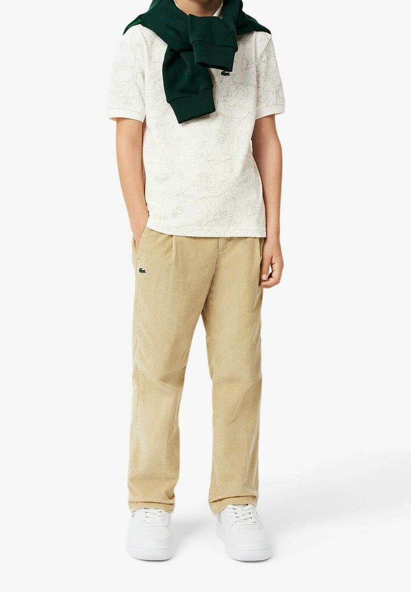 Enfant portant un pantalon beige en velours côtelé, un polo blanc à motif floral, des baskets blanches et un pull vert posé sur les épaules.