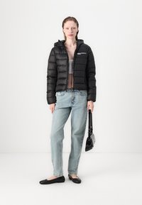 Juoda pūkinė striukė su gaubtu, horizontaliomis siūlėmis. Dėvima su šviesiai mėlynais džinsais, juodais lygiais batais ir maža juoda rankine.