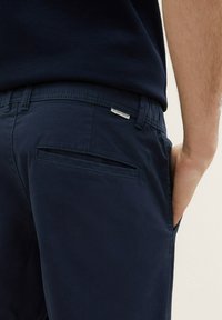 Detailní záběr na osobu v námořnických modrých kalhotách s zadní kapsou a ruku položenou v přední kapse.