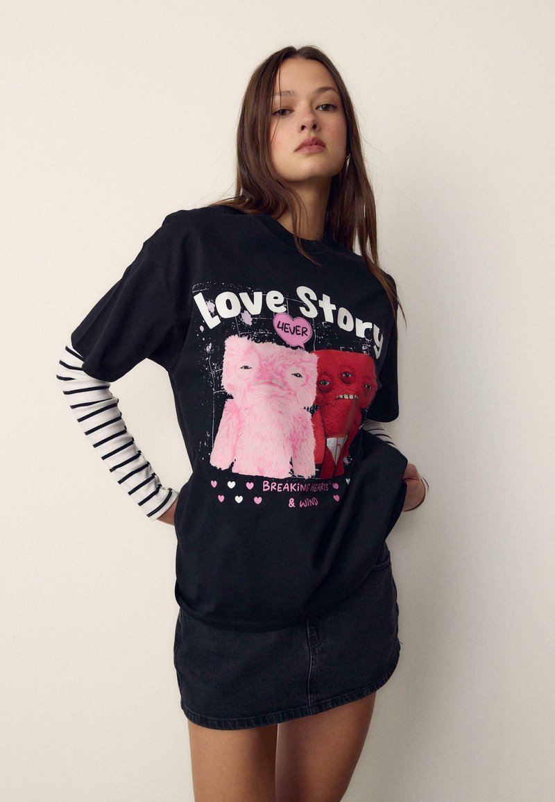 Jeune femme portant un t-shirt noir avec des créatures dessinées en rose et rouge, texte « Love Story 4EVER » et « Breaking Hearts & Wind », associé à des manches rayées et une jupe noire.