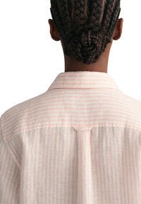 Hemd mit hellrosa und weißen Streifen, mit einem Kragen, einer Schulterpasse und einer mittleren Falte auf der Rückseite. Weicher, strukturierter Stoff.