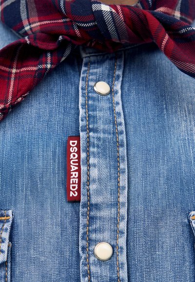 Close-up of blue denim shirt with metal snap buttons and red plaid scarf tied around the neck. Red brand tag reads DSQUARED2.